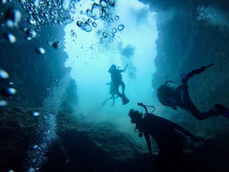 The image shows a group of scuba divers exploring an underwater cave. The divers are silhouetted against the bright light coming from the cave entrance. The water is clear and blue, and there are many bubbles rising to the surface. The cave walls are rocky and covered in marine life.